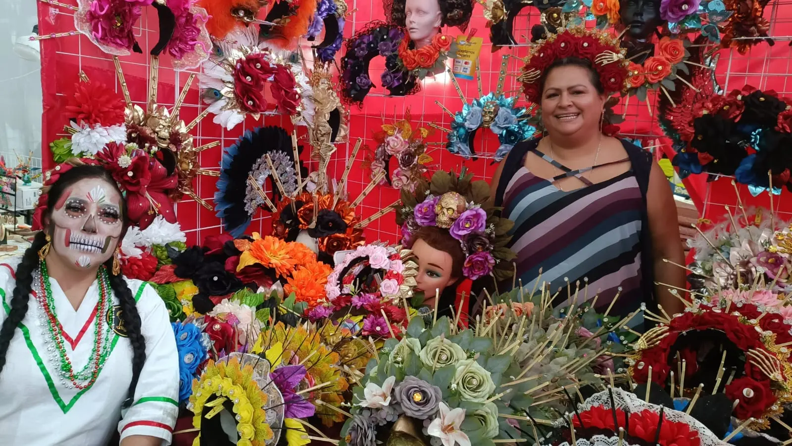 Puesto de tocados artesanales TocadosMon en un mercado de Día de Muertos, con una artesana y una clienta caracterizada de Catrina